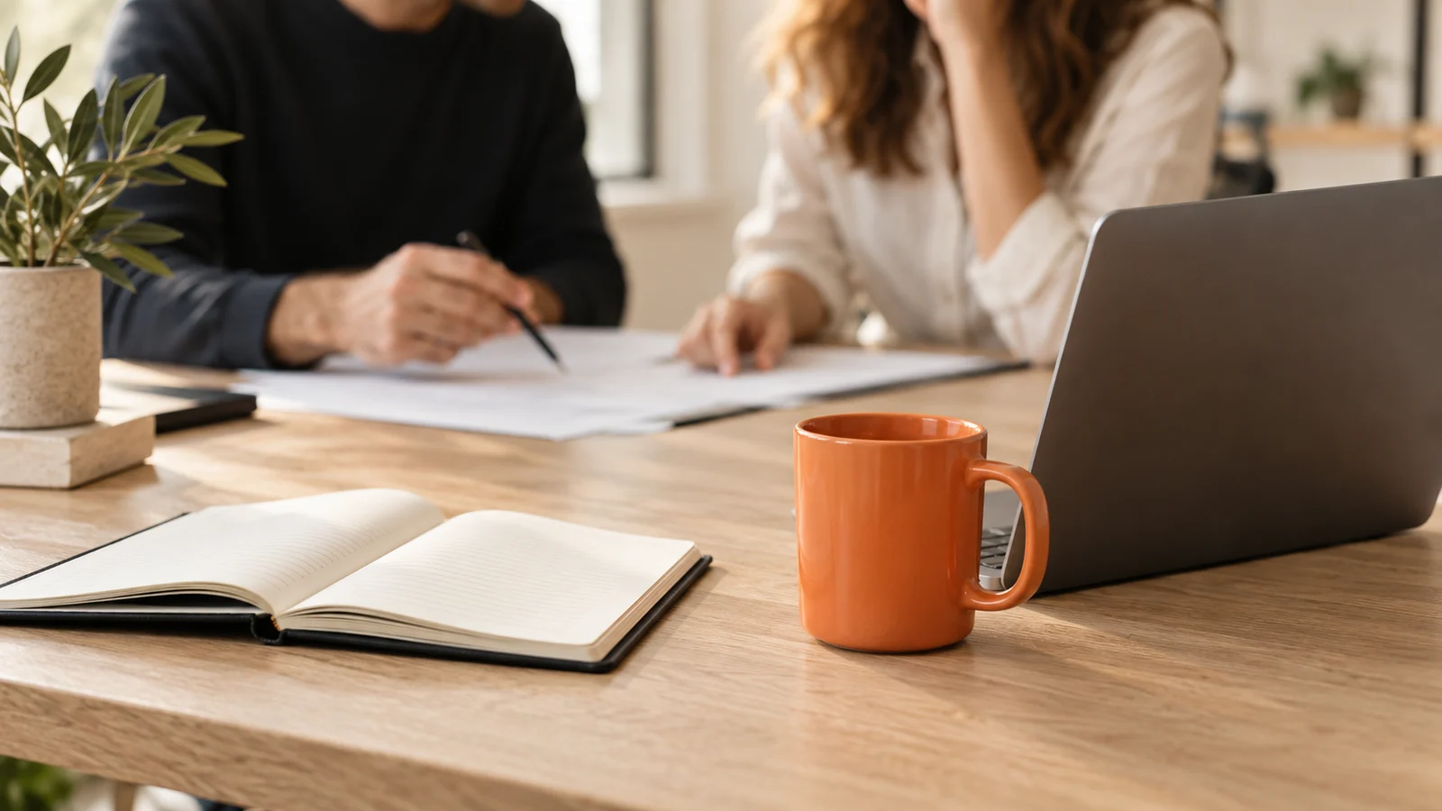 A charcoal laptop, cream notebook and orange mug in a collaborative business workspace